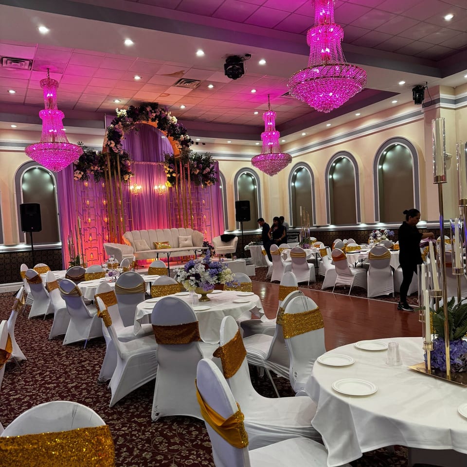 A large event hall with pink chandeliers, white tables, gold-accented chairs, and a decorated stage.